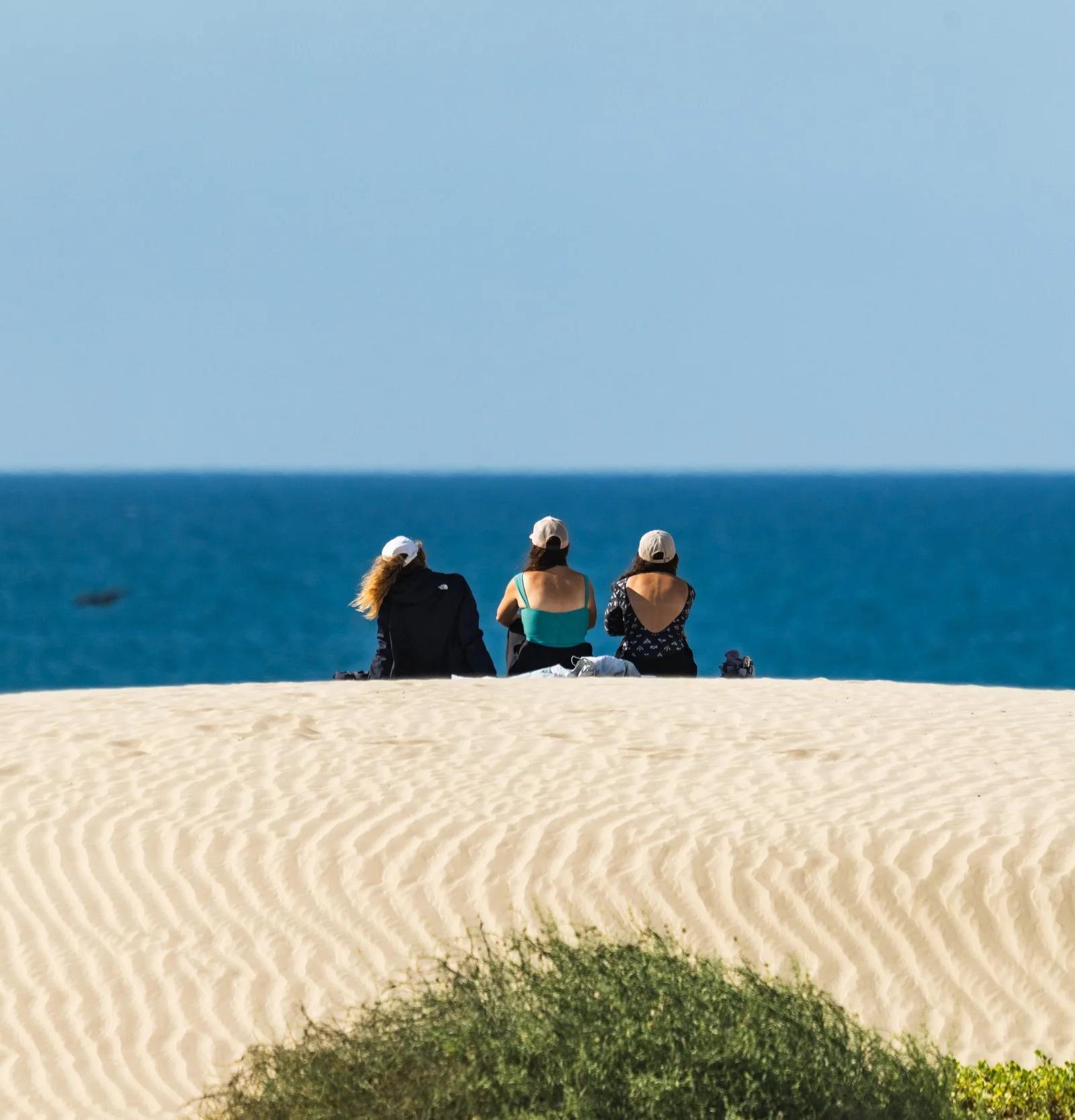 Tres viajeros sentados en lo alto de una duna de arena mirando el mar en Bahía Magdalena