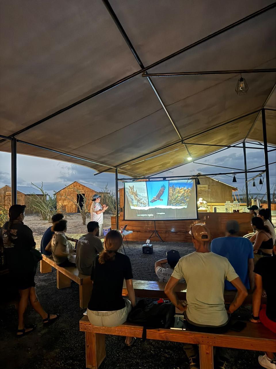 Un guía da una charla con proyector a un grupo sentado en bancas bajo una carpa al anochecer