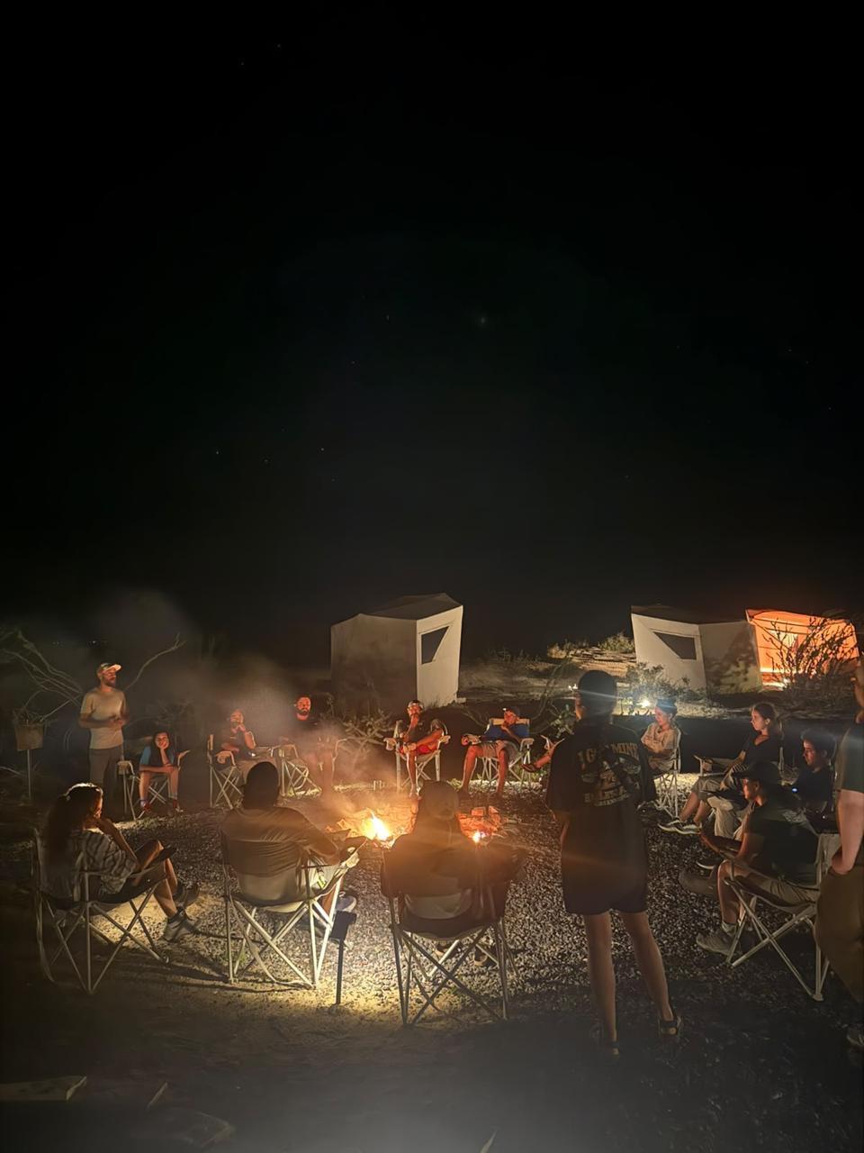 Huéspedes y guías sentados alrededor de una fogata de noche, con tiendas del campamento al fondo